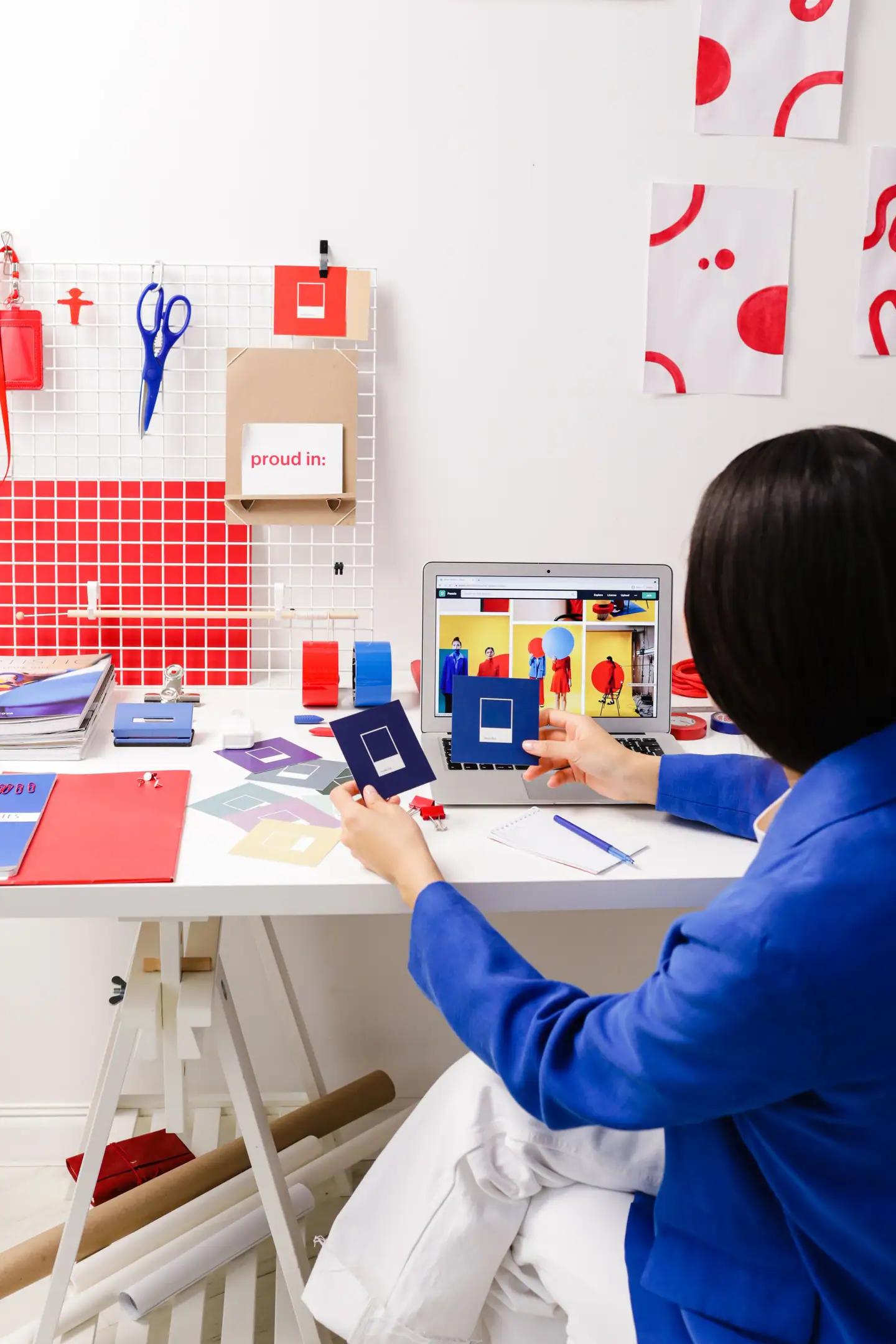 Designerin sortiert Farbmuster am Arbeitstisch, an der Wand ein Gitter mit Scheren und Entwürfen
