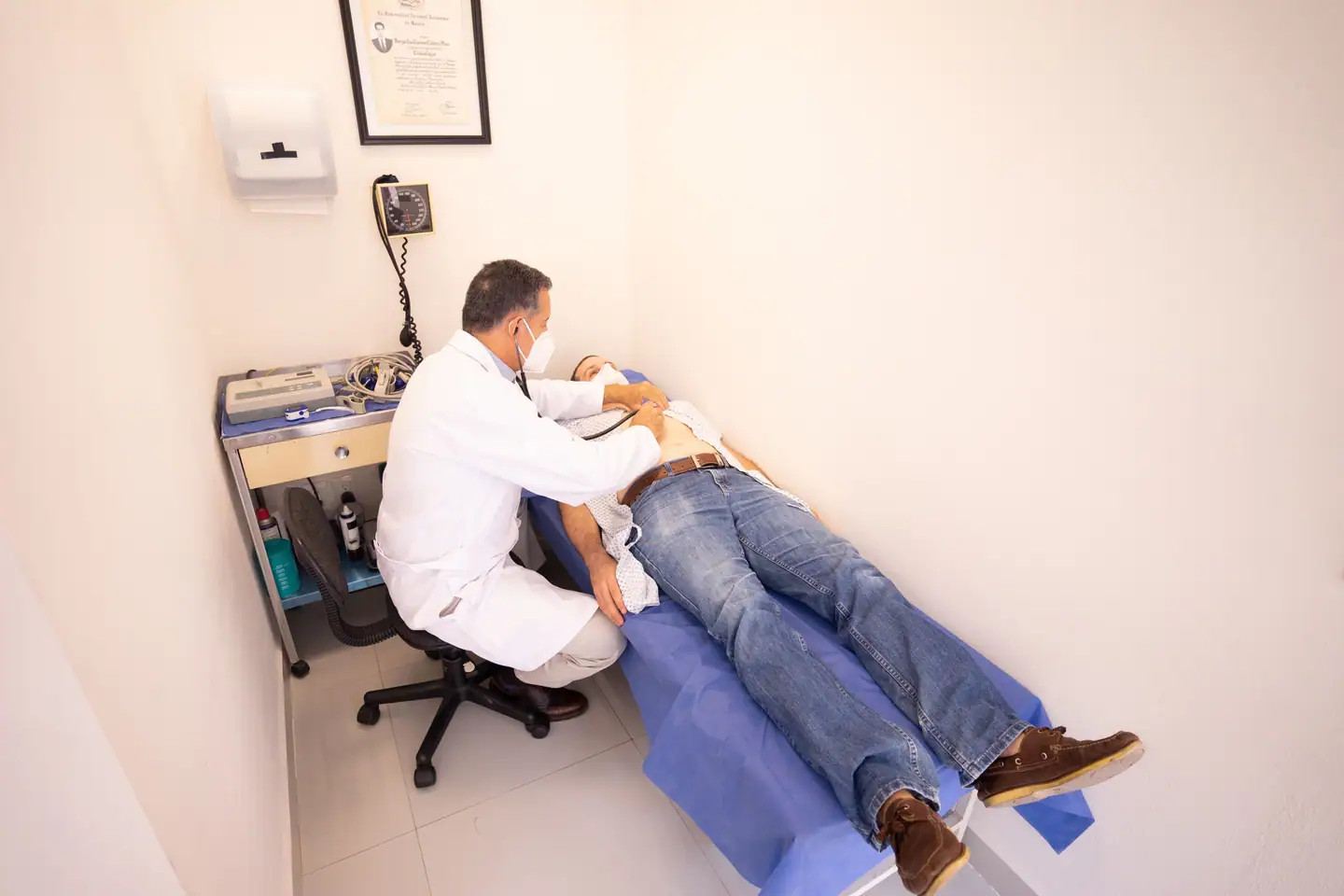 Arzt untersucht einen liegenden Patienten, Wandgerät und Diplom an der Wand