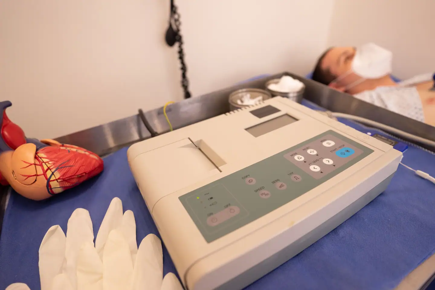 EKG-Gerät auf der Liege, daneben Handschuhe und ein Herzmodell
