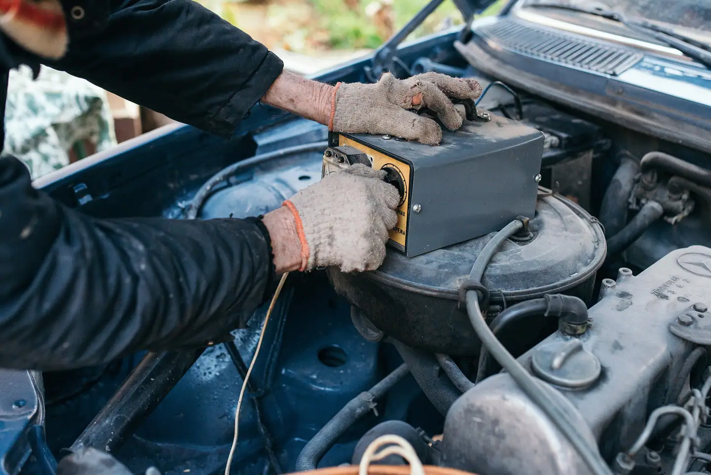 Hände in Arbeitshandschuhen messen die Batteriespannung im Motorraum
